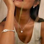A close-up lifestyle shot of a woman wearing a delicate silver pendant necklace with spiral texture and a knotted silver cuff bracelet, set against a soft beige background.