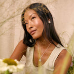 A woman with long dark hair wearing a silver pendant necklace and matching silver stud earring, posing in a light beige sleeveless top. The image has a soft, dreamy aesthetic with a double-exposure overlay of dried grass and flowers, and blurred green foreground foliage.