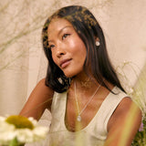 A woman with long dark hair wearing a silver pendant necklace and matching silver stud earring, posing in a light beige sleeveless top. The image has a soft, dreamy aesthetic with a double-exposure overlay of dried grass and flowers, and blurred green foreground foliage.