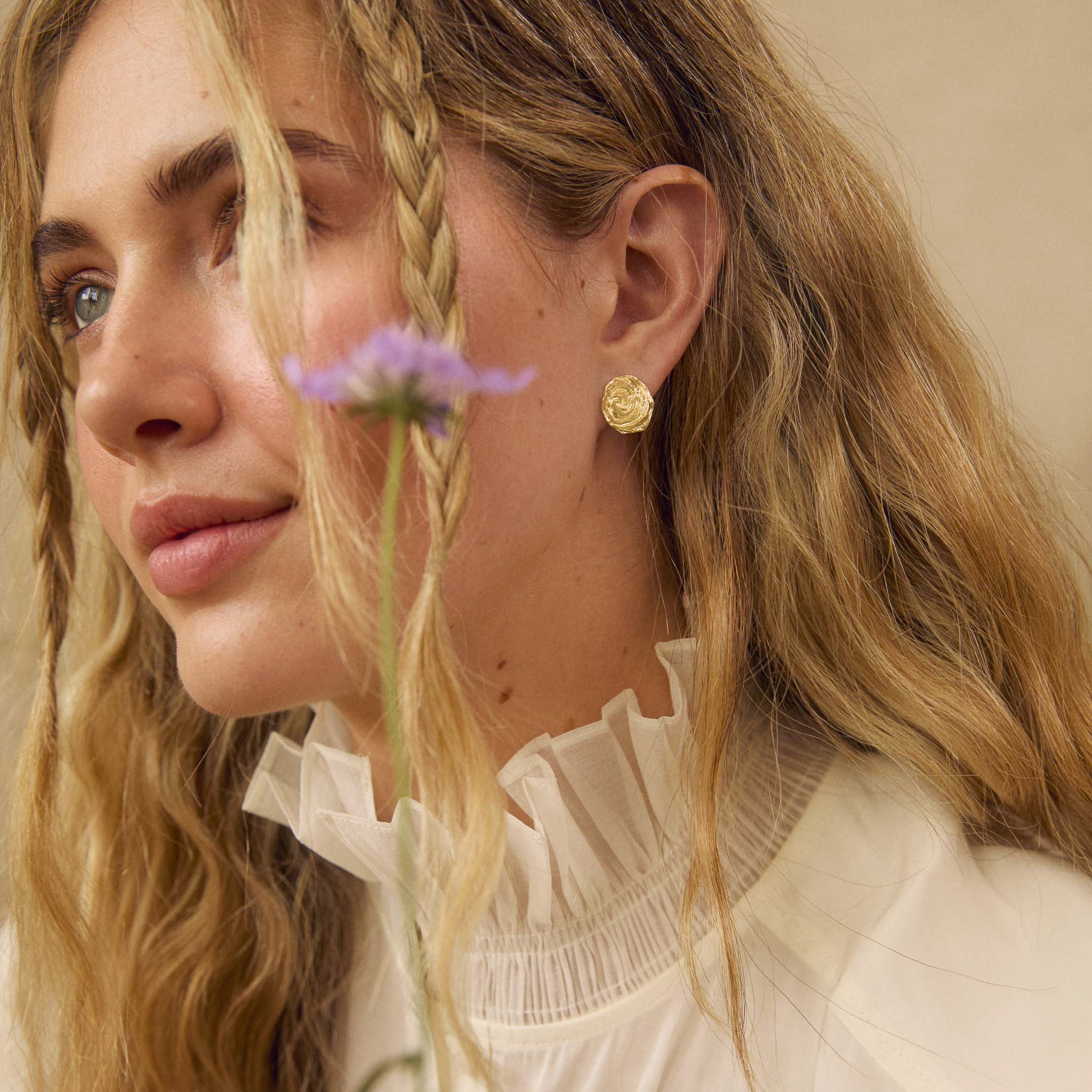 A close-up lifestyle shot of a woman with blonde hair wearing a white ruffled blouse and a textured gold stud earring, with a small purple flower resting against her face.