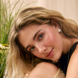 A close-up lifestyle portrait of a young woman with blonde hair, wearing a beige skirt, black top and silver spiral stud earrings, looking directly at the camera with a soft smile.