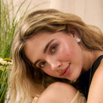 A close-up lifestyle portrait of a young woman with blonde hair, wearing a beige skirt, black top and silver spiral stud earrings, looking directly at the camera with a soft smile.