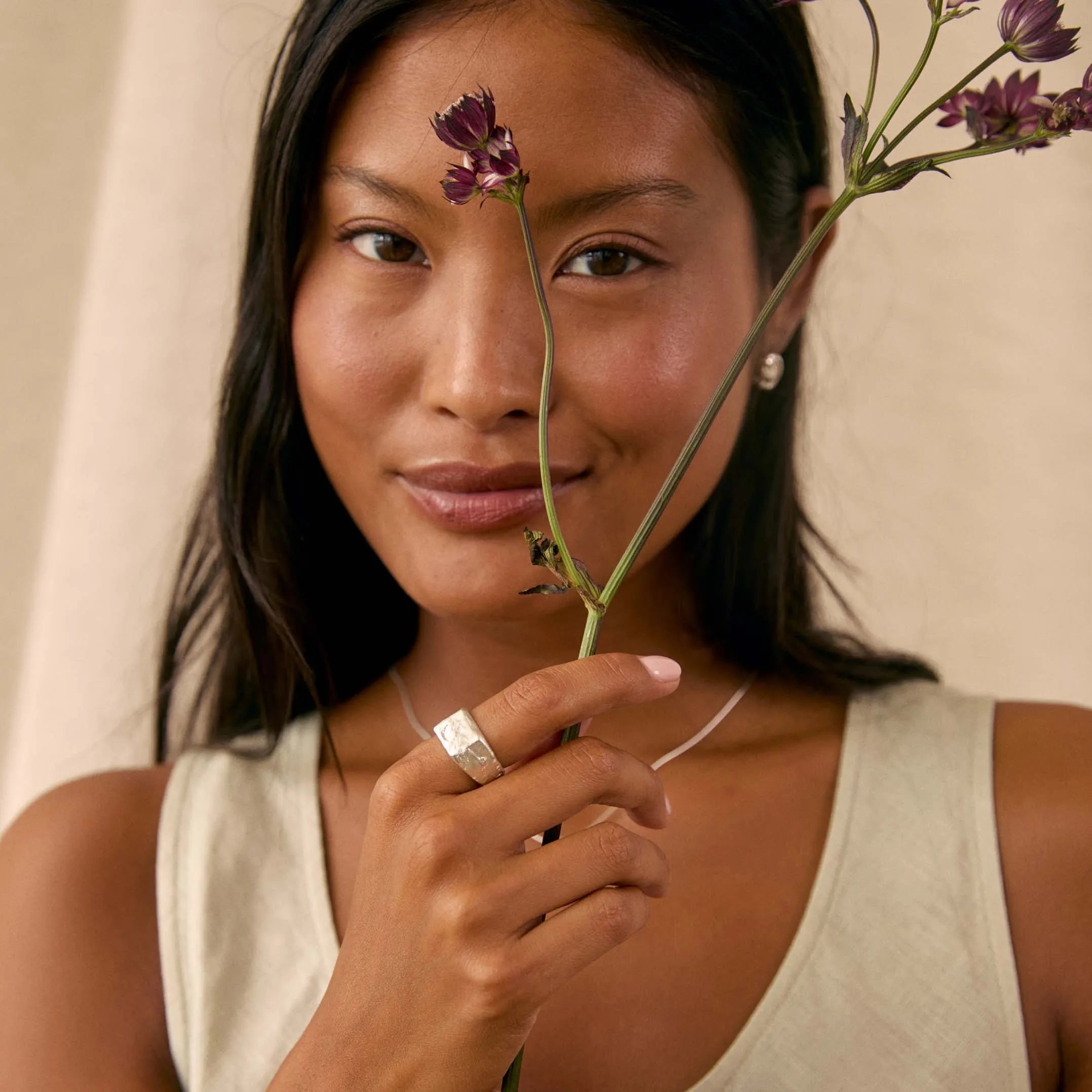 A woman wearing a silver ring and silver bracelets holds a delicate purple flower stem in front of her face, wearing a beige sleeveless top against a neutral beige background.