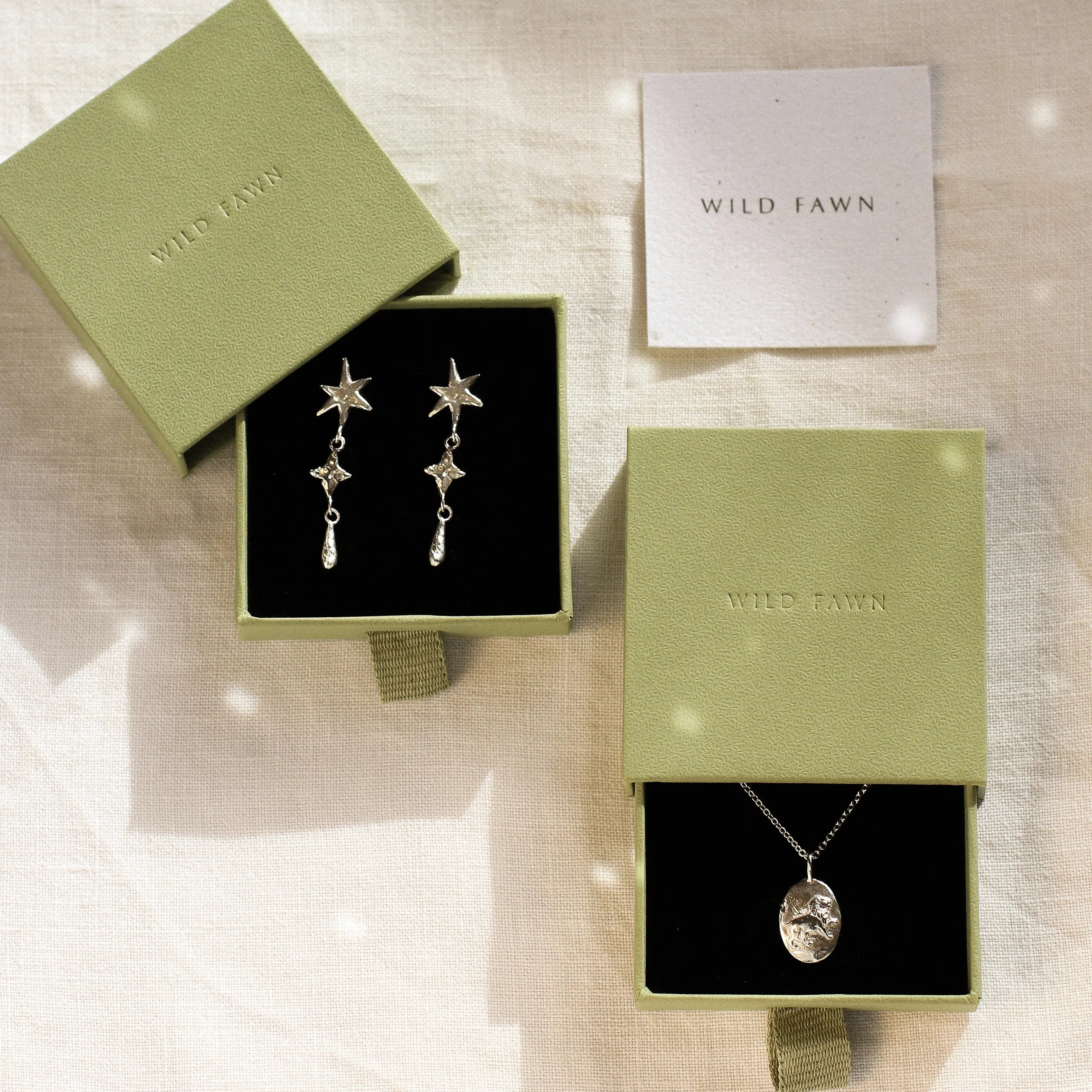 Jewelry set with earrings and a leo necklace in 'Wild Fawn' green boxes on a textured surface.