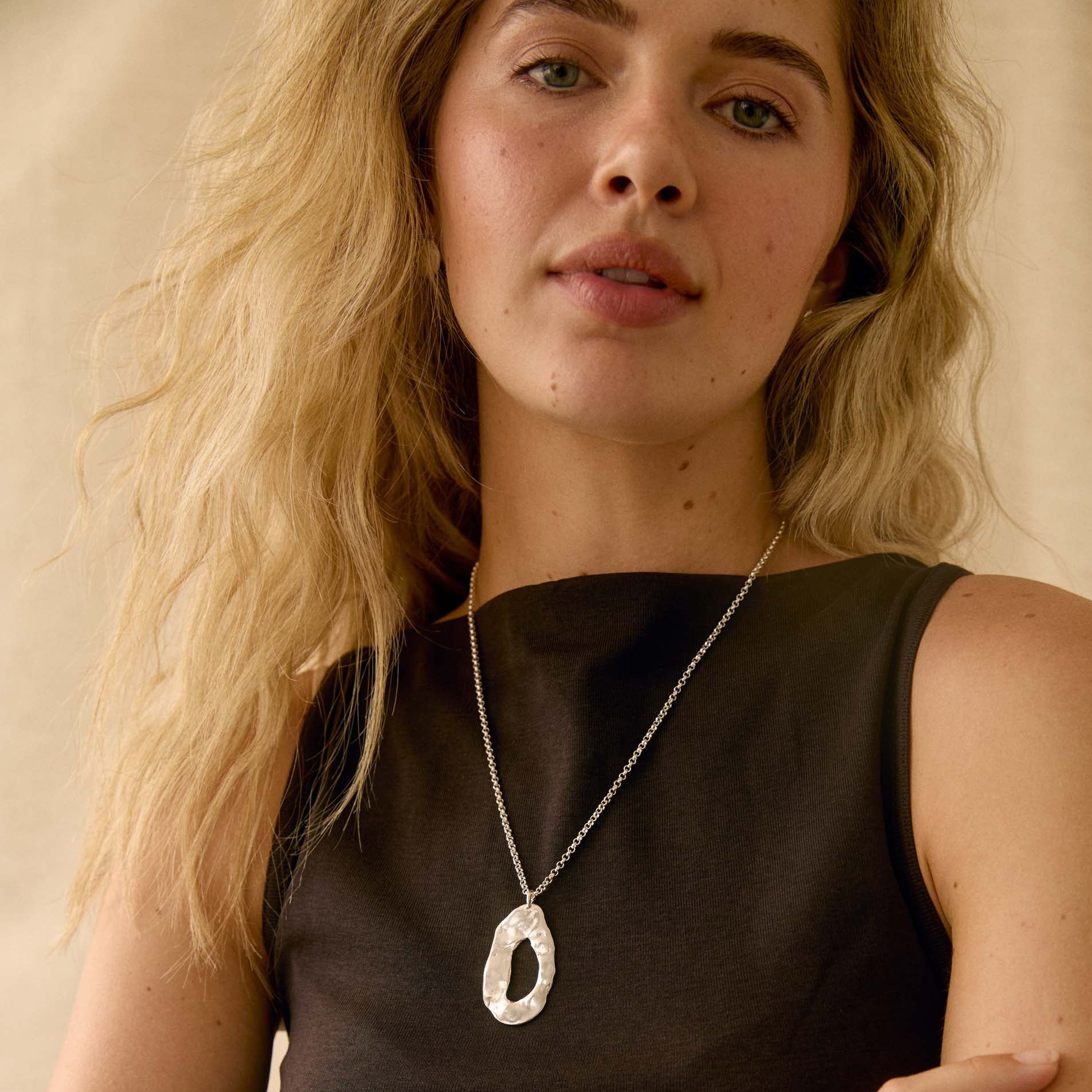 A woman wearing a silver pendant necklace with a textured, organic oval shape. She is wearing a black sleeveless top.
