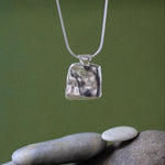 A silver pendant necklace with a textured, hammered square pendant hanging over a stack of smooth gray river stones against a soft green background.