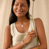A smiling woman wearing a silver geometric pendant necklace, silver stud earrings, and a silver ring, holding a purple flower stem against a neutral beige fabric backdrop.