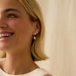 Smiling woman wearing diamond sun and star earrings, a white sleeveless wedding dress with a plain background