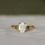 Gold diamond engagement ring with a oval diamond and round green tourmaline gemstones on a textured surface