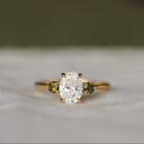 Gold diamond engagement ring with a oval diamond and round green tourmaline gemstones on a textured surface