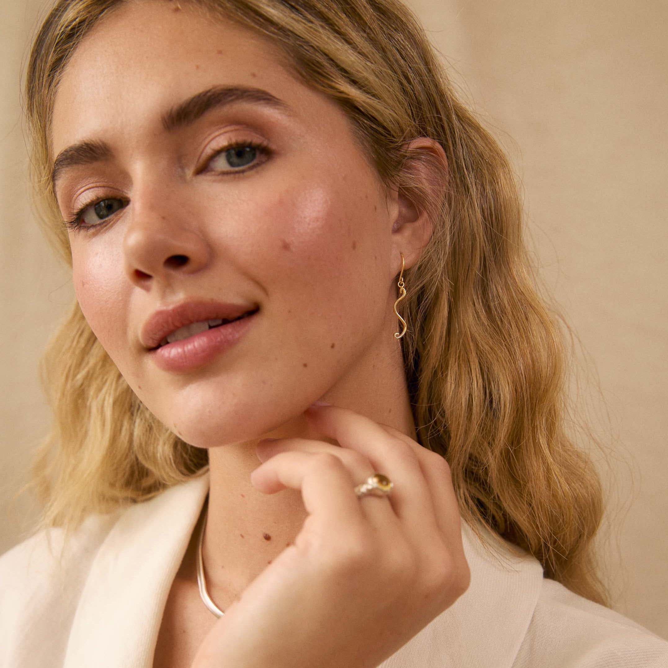 A close-up lifestyle shot of a woman with blonde hair wearing a delicate gold ring on her finger and a small dangle earring, dressed in a white blazer against a warm beige background.