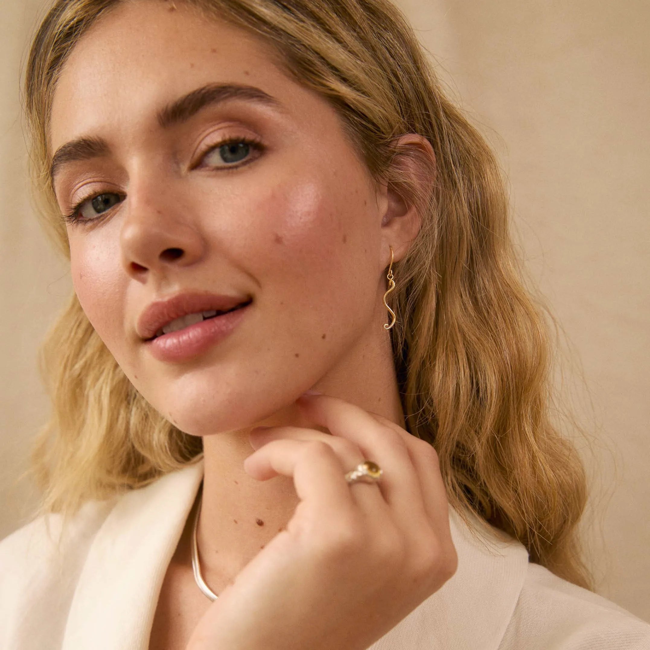 A close-up lifestyle shot of a woman with blonde hair wearing a delicate gold ring on her finger and a small dangle earring, dressed in a white blazer against a warm beige background.