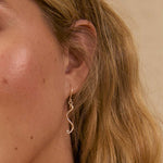 close up of a blonde womans ear wearing a delicate thin silver dangle earring with s-shaped pendant