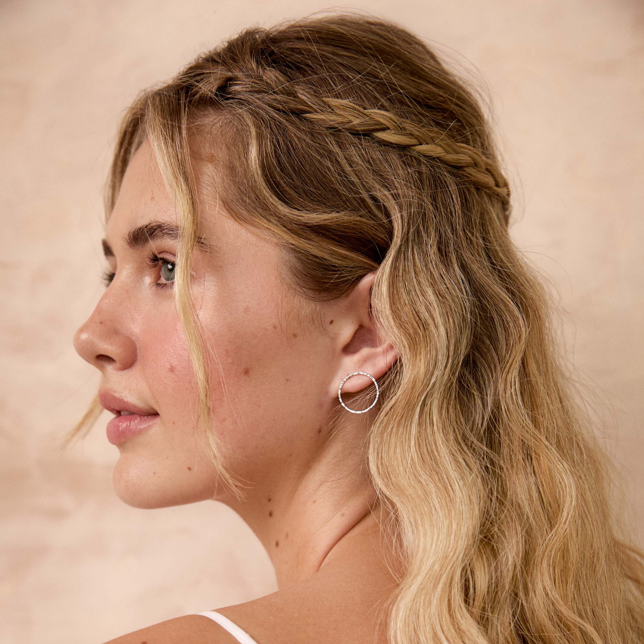 A woman shown in side profile wearing a delicate silver hoop earring, with long blonde wavy hair and a braided detail across the top of her head, against a warm beige background.