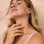 A blonde woman with her eyes closed, wearing a white spaghetti-strap top, gold hoop earrings and a wavy gold ring, posing against a textured beige wall.
