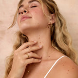 A blonde woman with her eyes closed, wearing a white spaghetti-strap top, gold hoop earrings and a wavy gold ring, posing against a textured beige wall.