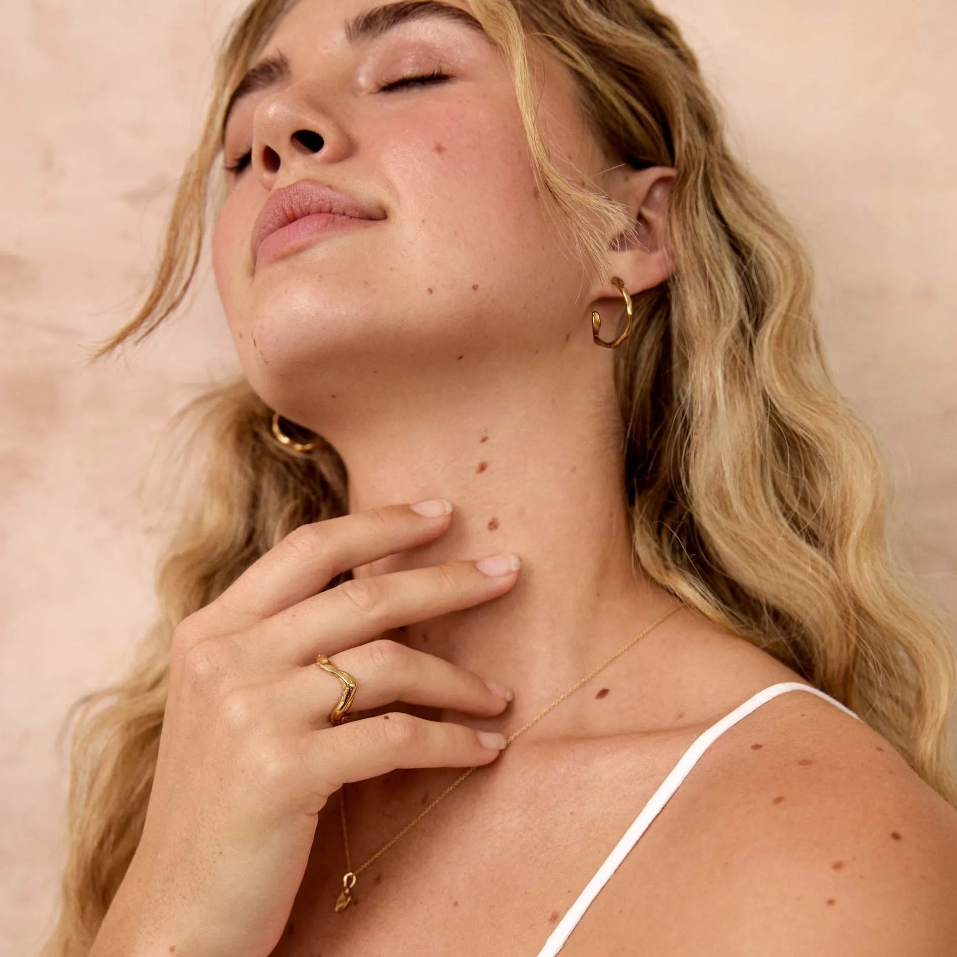 A blonde woman with her eyes closed, wearing a white spaghetti-strap top, gold hoop earrings and a wavy gold ring, posing against a textured beige wall.