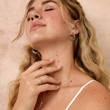 A blonde woman with her eyes closed, wearing a white spaghetti-strap top and delicate silver jewelry, including a necklace and hoop earrings, posing against a textured beige wall.