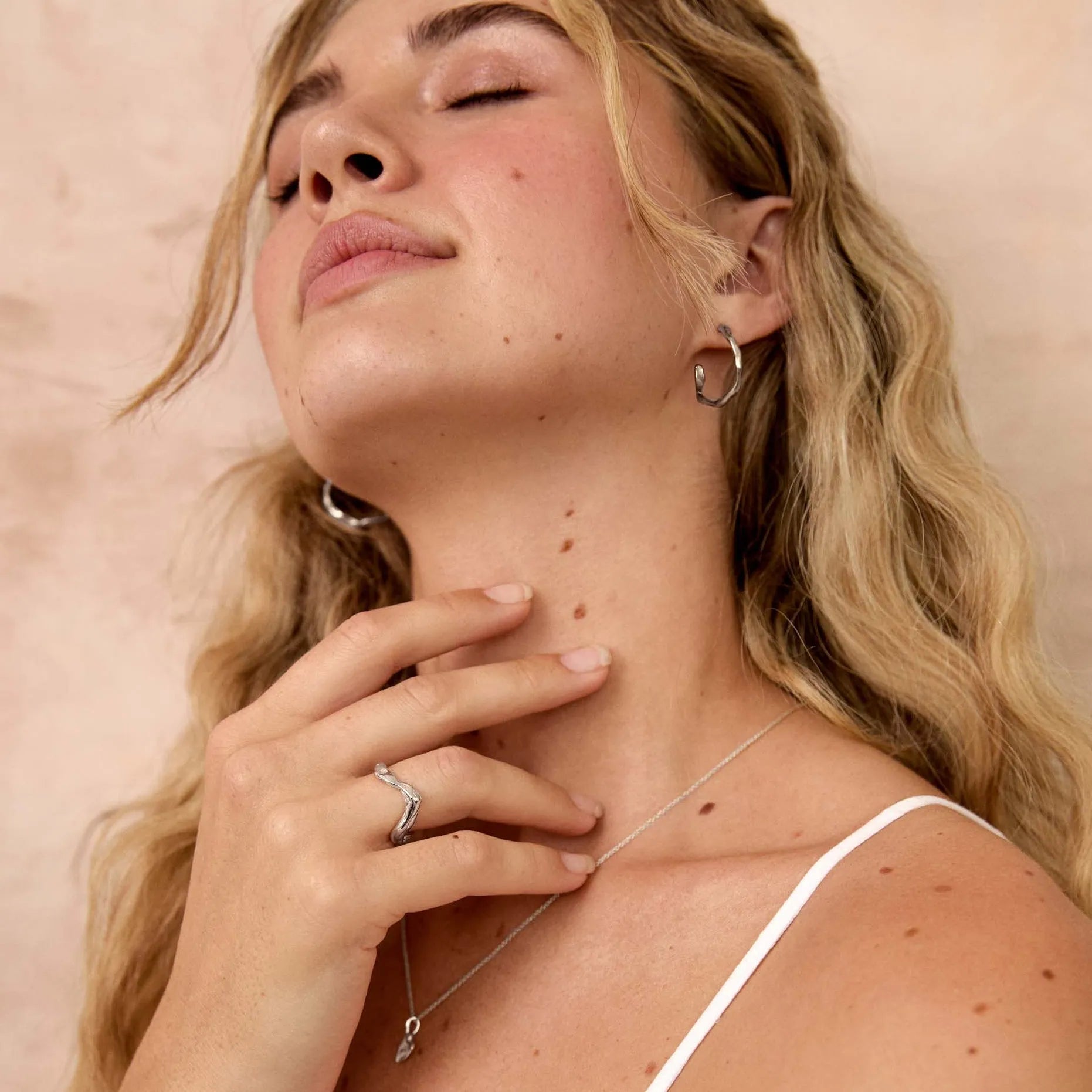 A blonde woman with her eyes closed, wearing a white spaghetti-strap top and delicate silver jewelry, including a necklace and hoop earrings, posing against a textured beige wall.