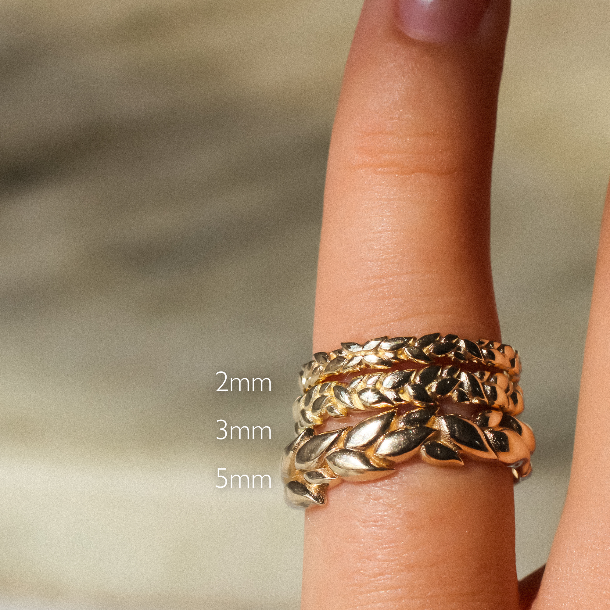 A close-up of a gold leaf-patterned wrap ring worn on a finger, with size text overlays indicating 2mm, 3mm, and 5mm widths against a soft blurred beige background.