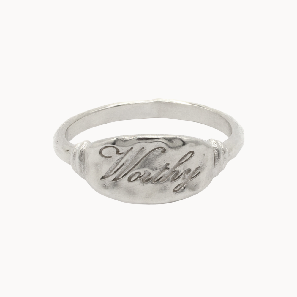 A silver signet ring with a textured oval face featuring the engraved script word 'Worthy', shown on a plain white background.
