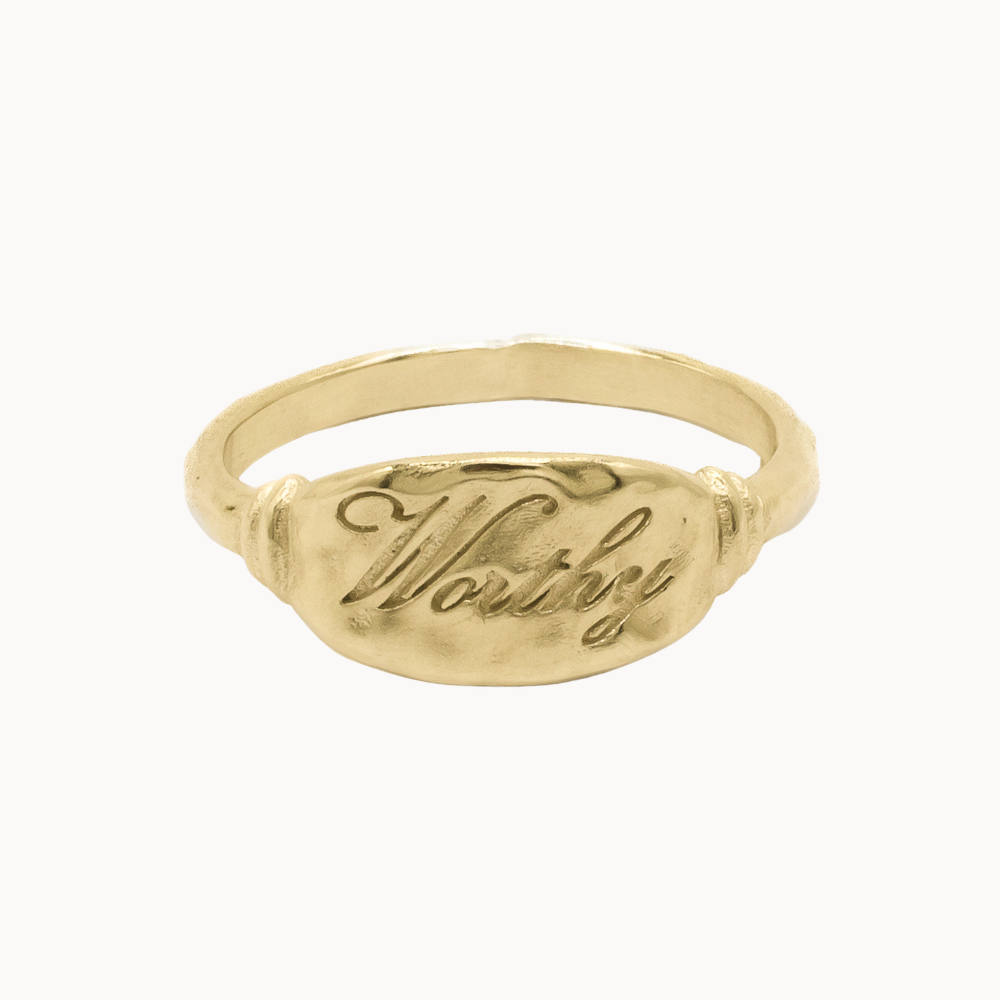 A gold signet ring with the word 'Worthy' engraved on the flat oval face, shown against a plain white background.