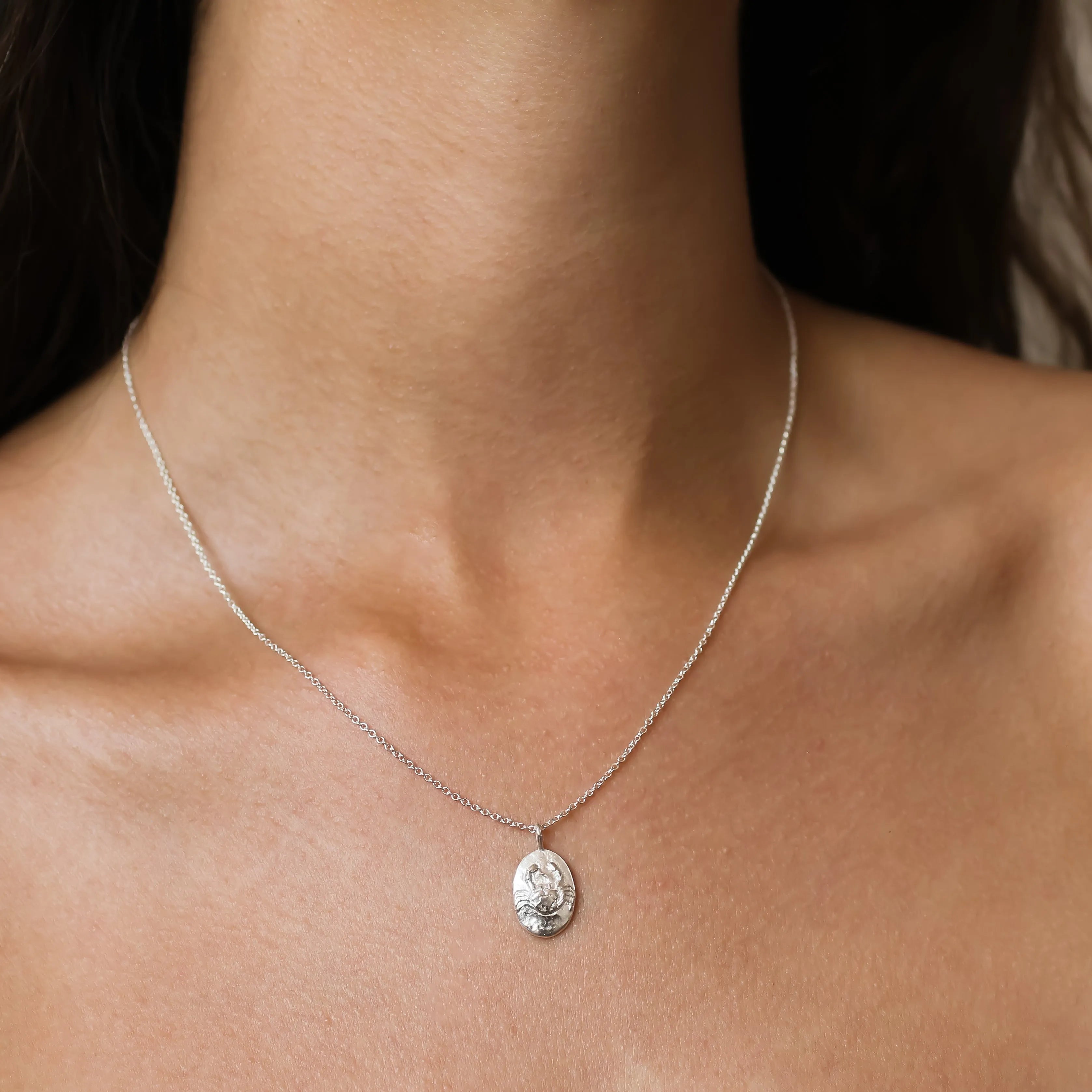 A close-up of a delicate silver chain necklace with an oval pendant featuring an embossed Cancer crab design, worn on a model's neck.