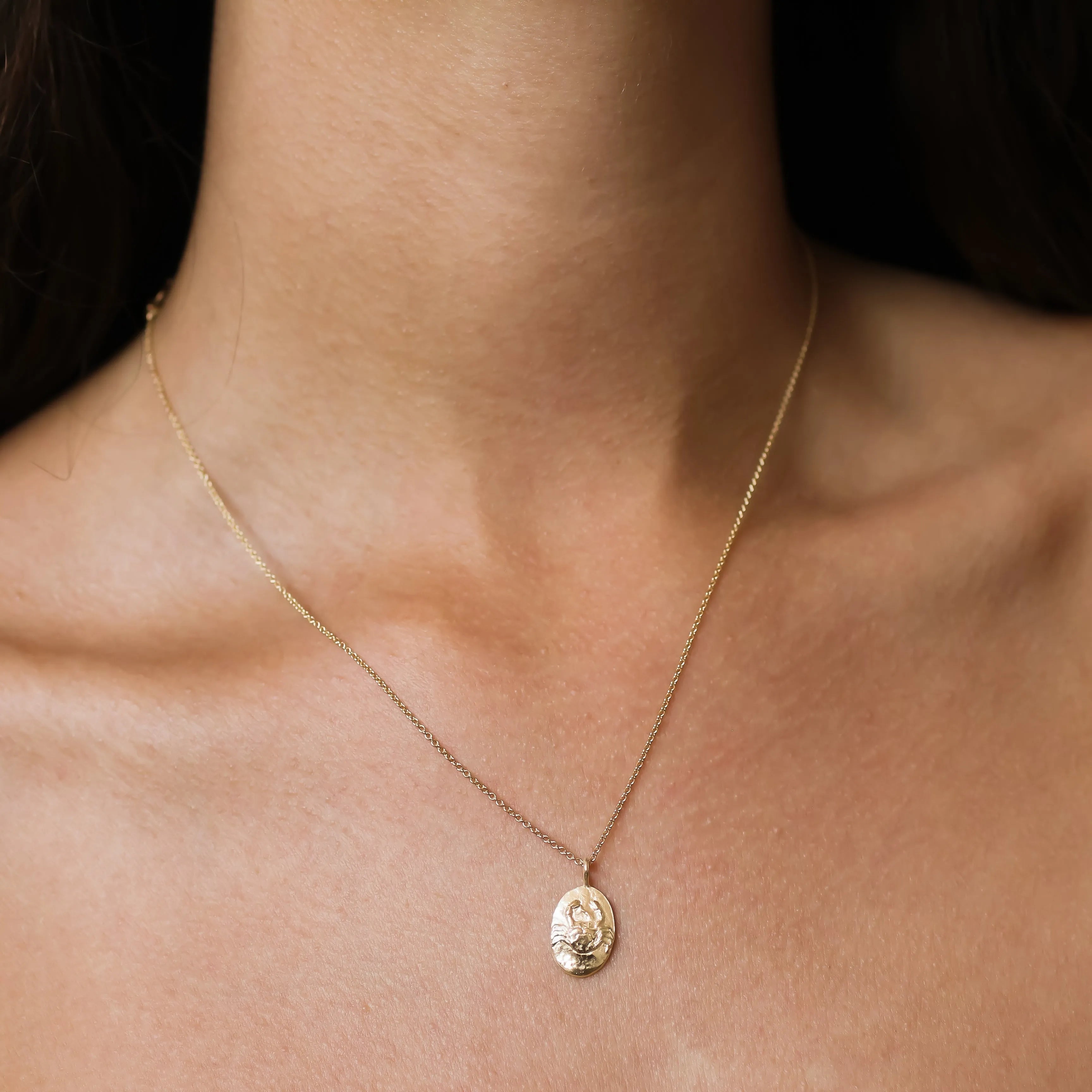 A delicate gold chain cancer zodiac necklace with an oval pendant featuring a raised classical-style face, worn on a model's neck.