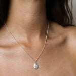 A close-up of a delicate silver chain necklace with an oval, textured silver capricorn pendant, worn on a model's neck.