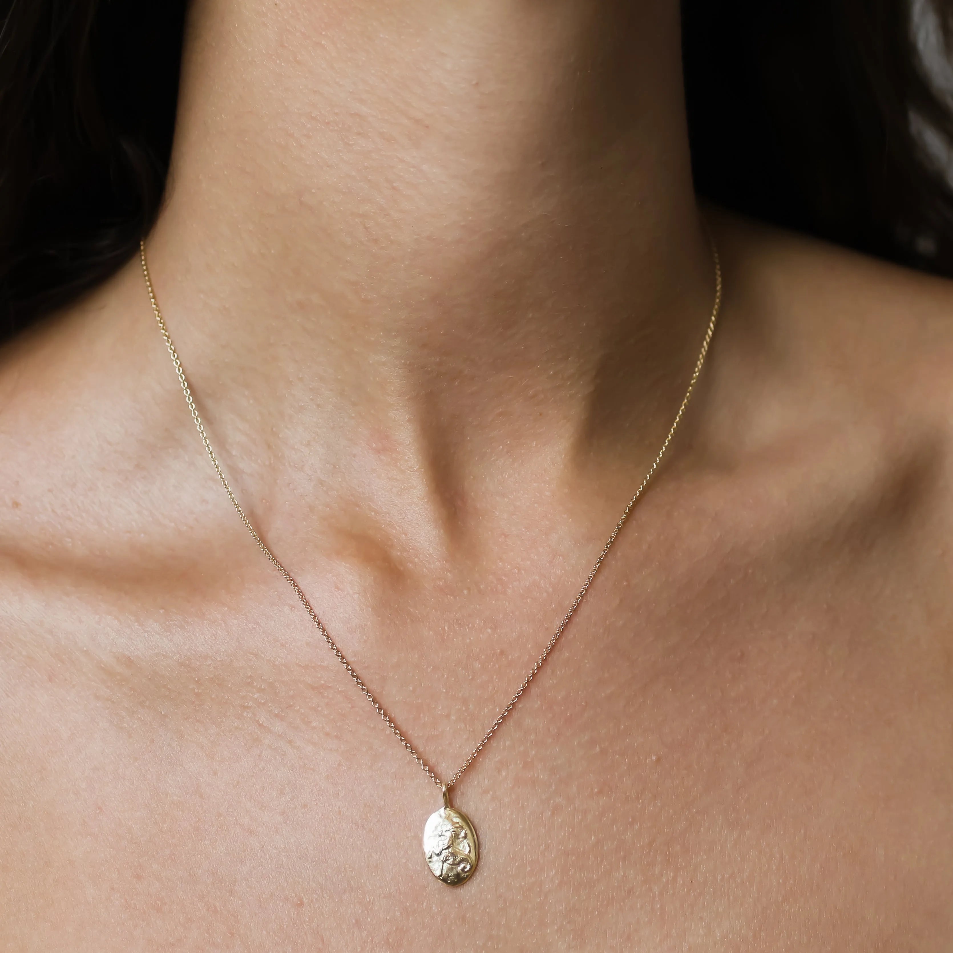 A close-up of a person's neck and upper chest wearing a delicate gold Capricorn zodiac necklace with an oval carved pendant.