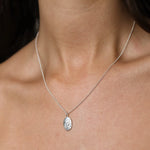 A close-up of a person's neck and chest wearing a delicate silver chain necklace with an oval silver pendant featuring a Gemini design.