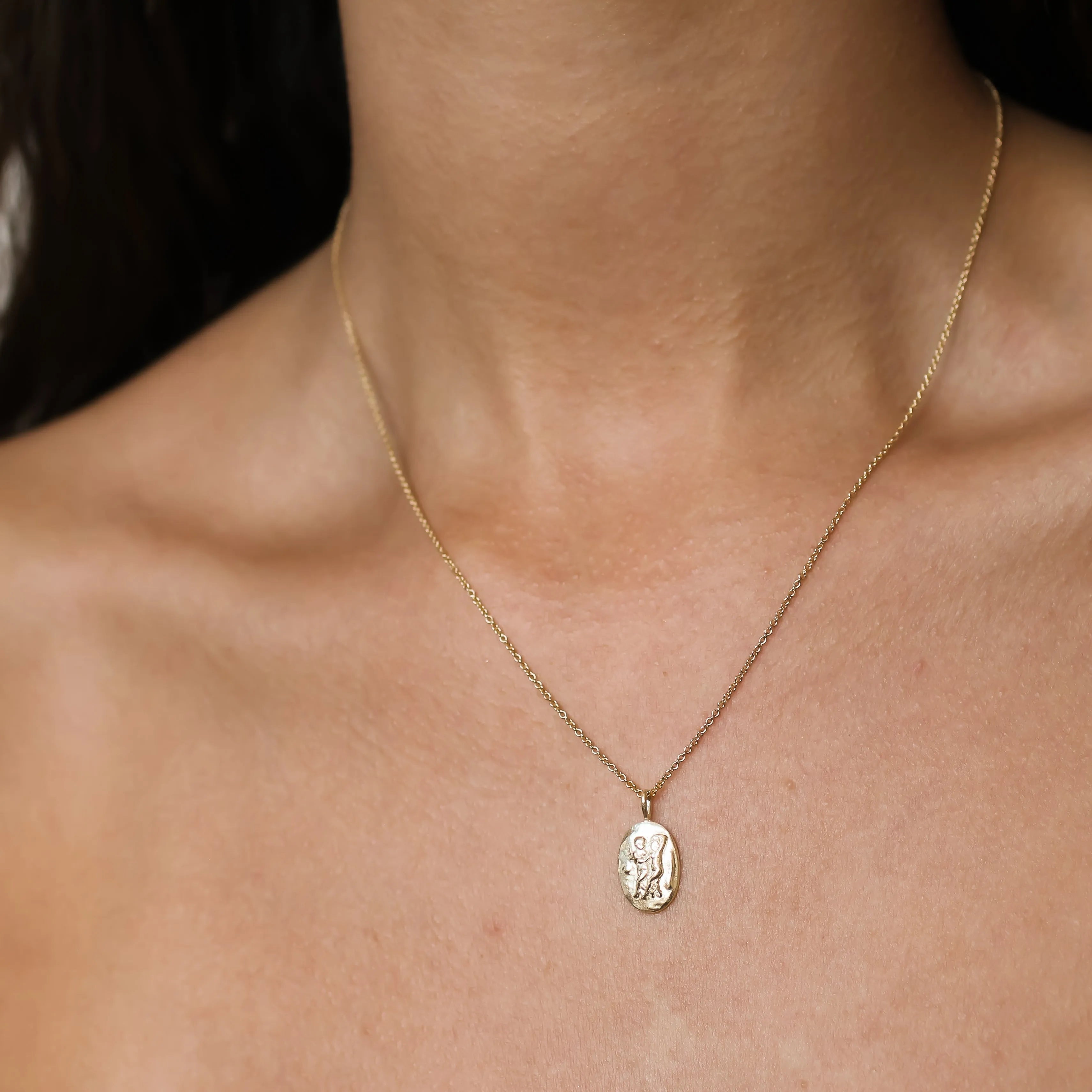 A delicate gold chain necklace with an oval pendant featuring an embossed Gemini symbol, shown worn on a person's neck and upper chest.