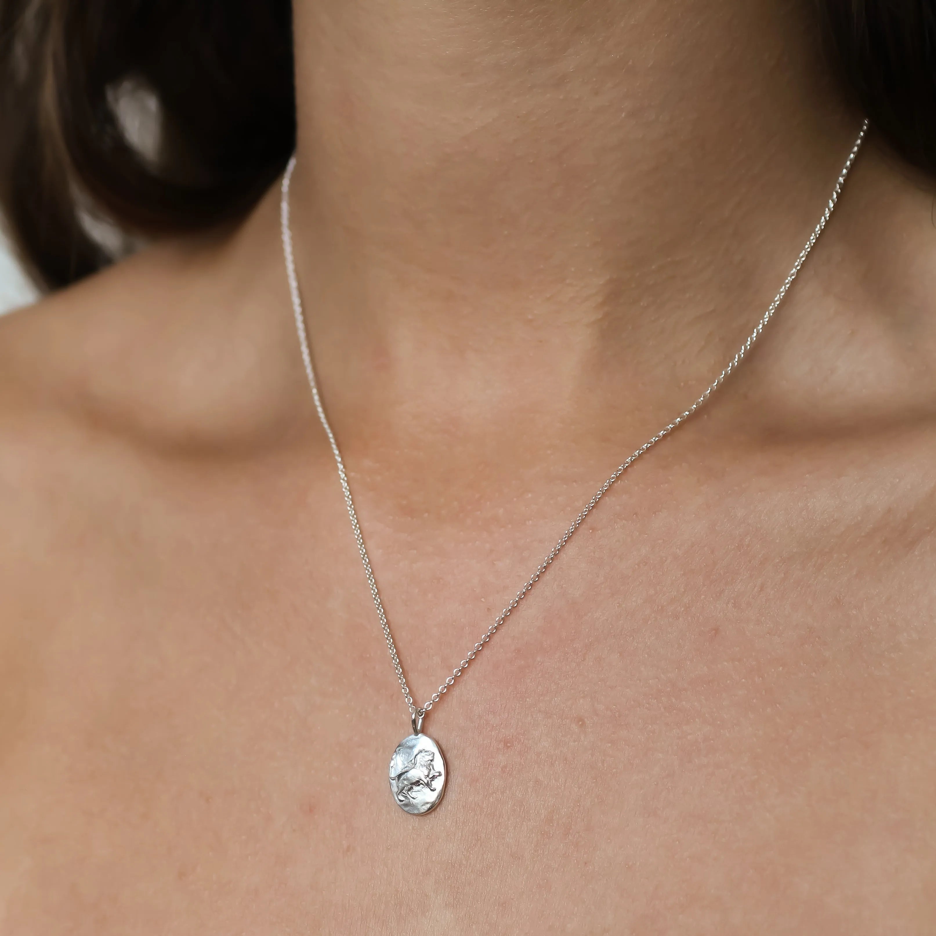 A close-up of a person's neck and upper chest wearing a delicate silver chain necklace with an oval pendant featuring a textured, Leo lion design/.