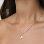 A close-up of a model's neck and chest wearing a delicate gold chain necklace with an oval pendant featuring an embossed Leo lion design.