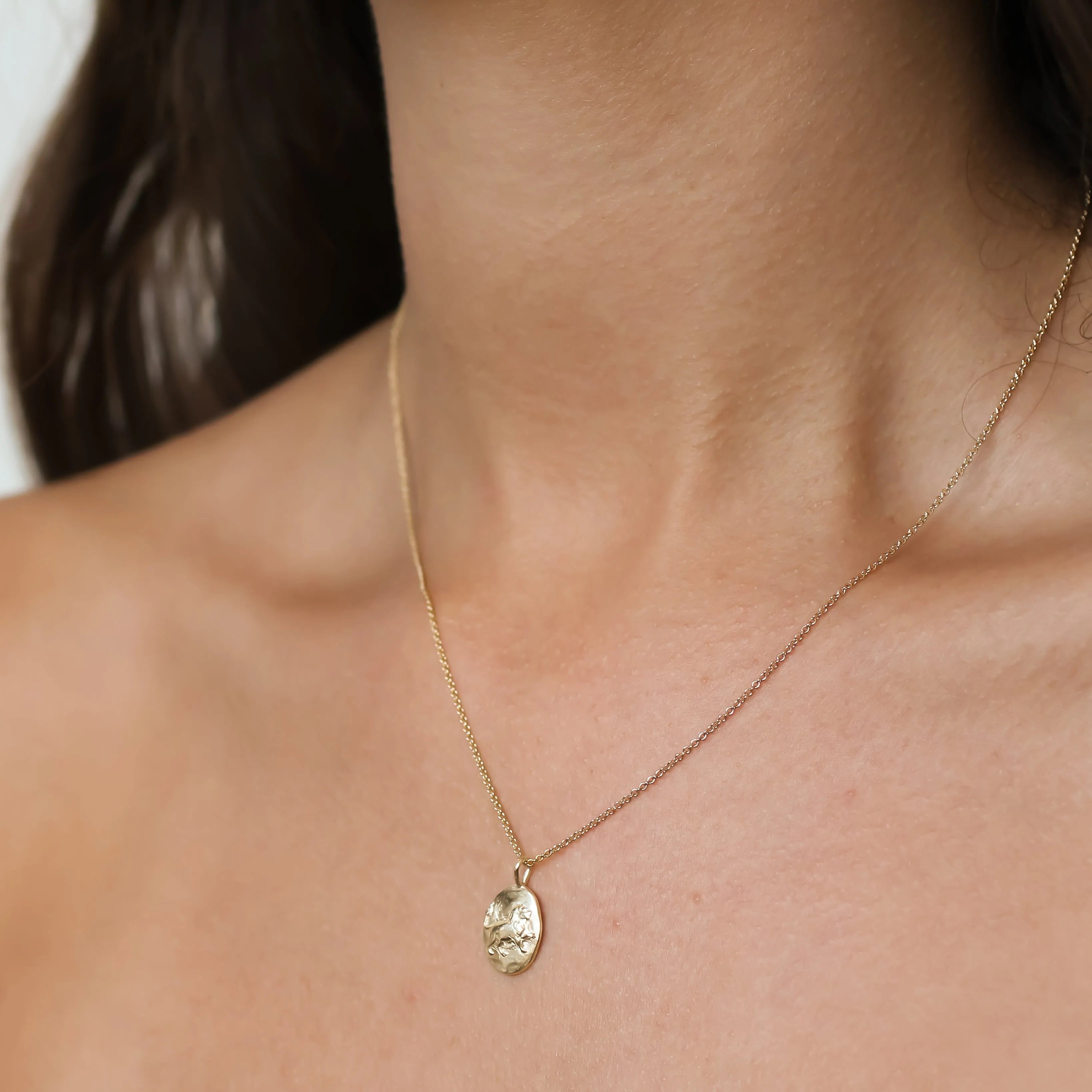A close-up of a model's neck and chest wearing a delicate gold chain necklace with an oval pendant featuring an embossed Leo lion design.
