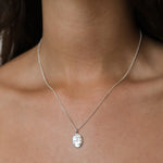 A close-up of a delicate silver chain Libra necklace with a small, oval, textured silver pendant worn on a model's neck.