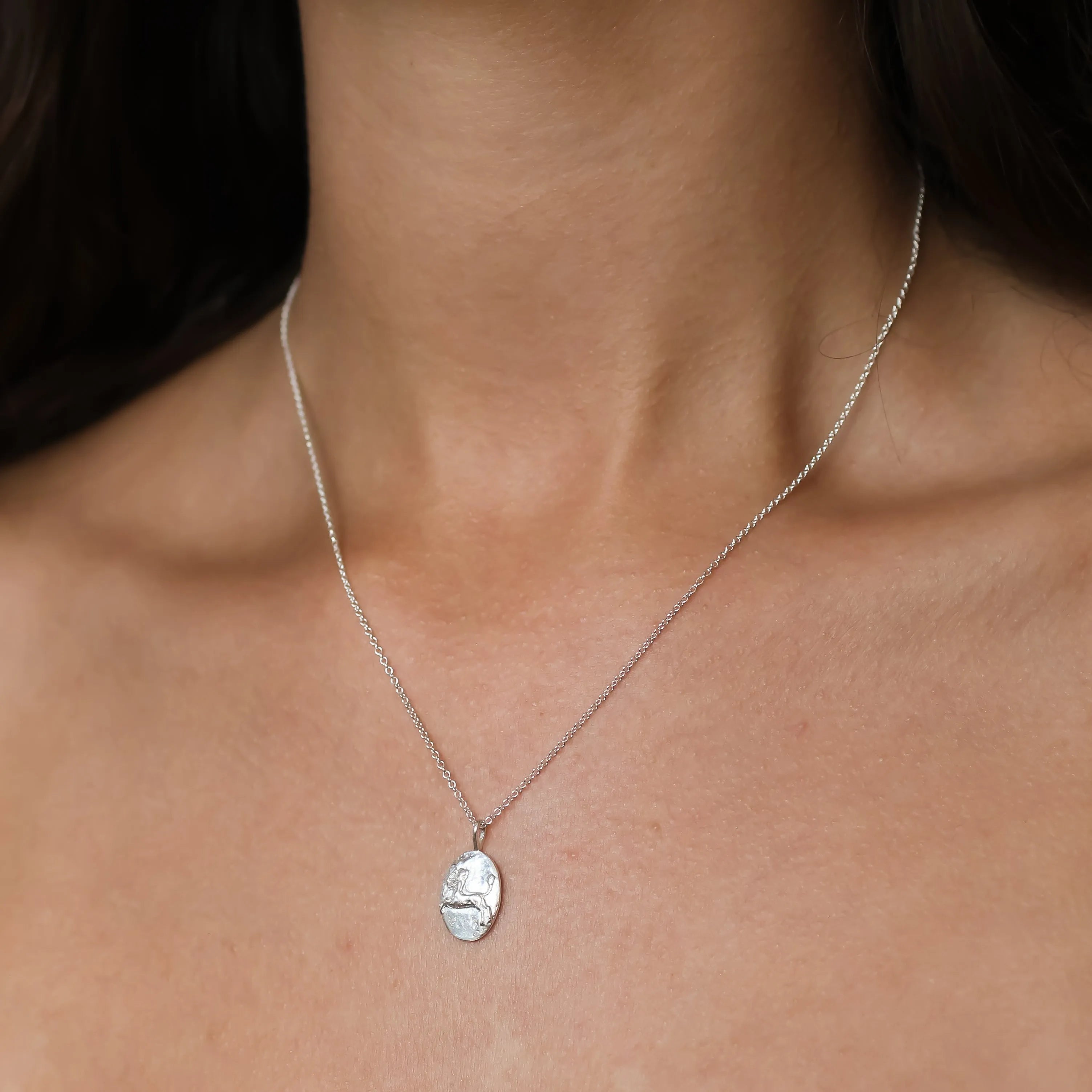 A close-up of a model's neck and chest wearing a delicate silver chain necklace with an oval textured sagittarius pendant.