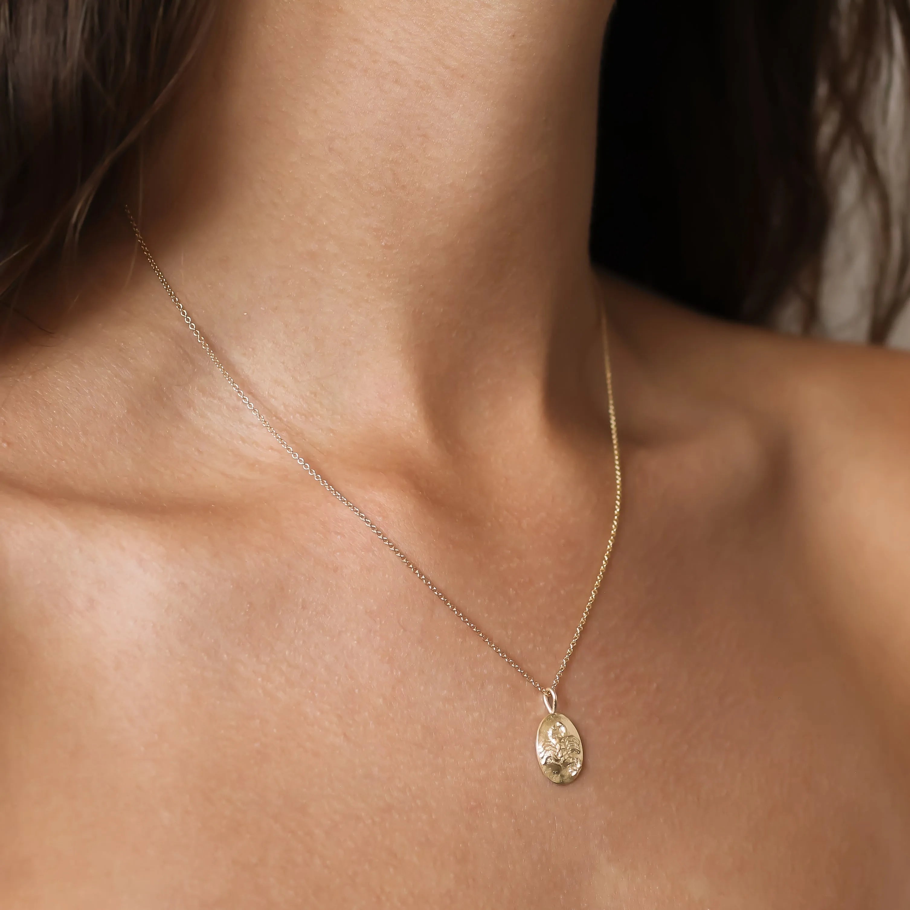A delicate gold chain zodiac necklace with an oval engraved Scorpio pendant worn on a model's neck, shown in a close-up shot.