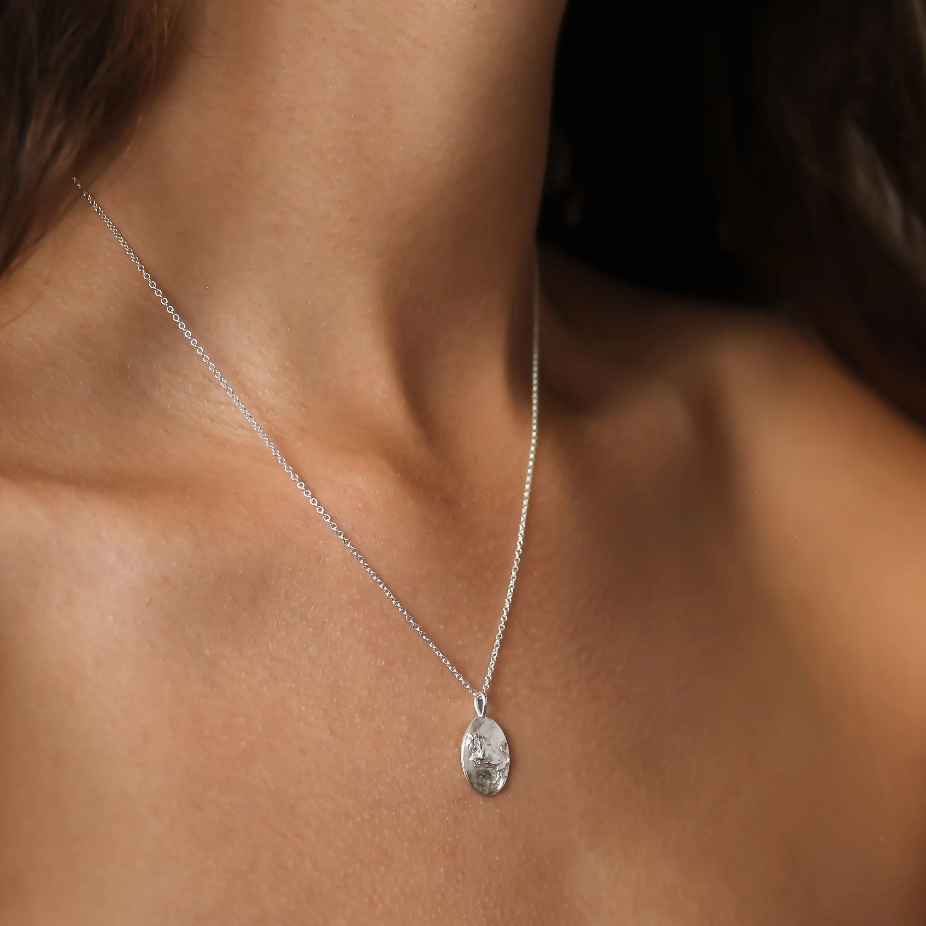 A close-up of a delicate silver chain necklace with an oval textured Taurus pendant worn on a model's neck.
