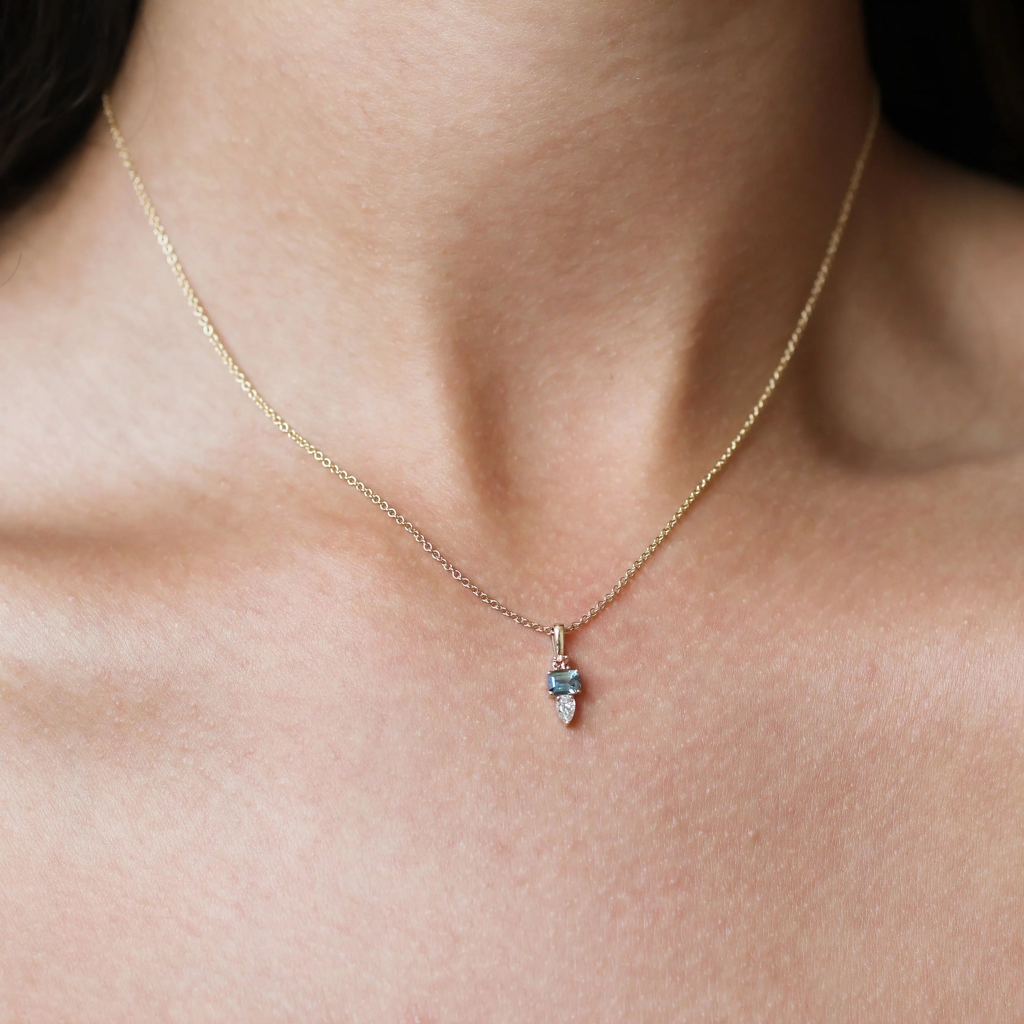 A delicate gold chain necklace with a small teardrop-shaped pendant featuring a peachy pink sapphire and light blue gemstone, shown worn on a model's neck.