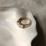 A gold three-stone ring with round diamonds and thick gold band resting on a textured ceramic dish against a neutral linen background.