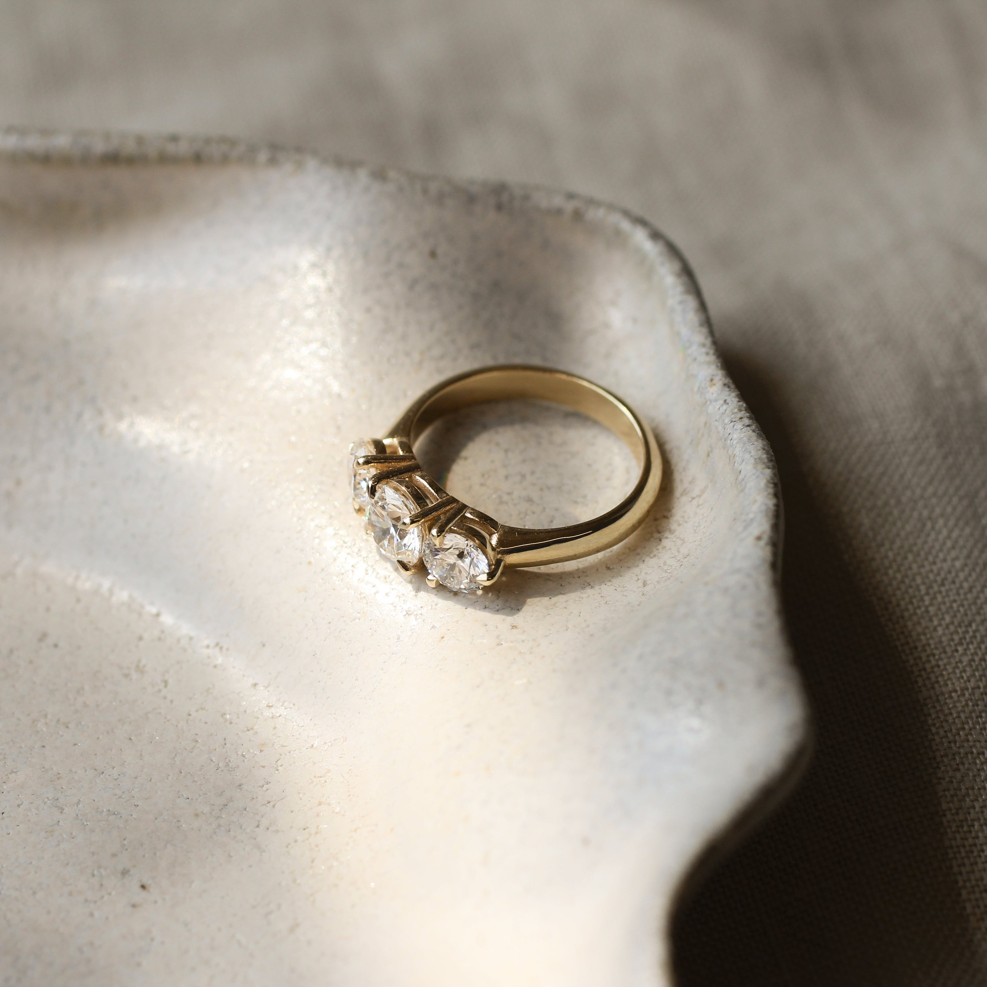 A gold three-stone ring with round diamonds and thick gold band resting on a textured ceramic dish against a neutral linen background.