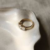 A gold three-stone ring with round diamonds and thick gold band resting on a textured ceramic dish against a neutral linen background.