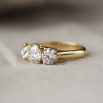 A yellow gold three-stone ring with round-cut clear gemstones, resting on a soft beige fabric surface.
