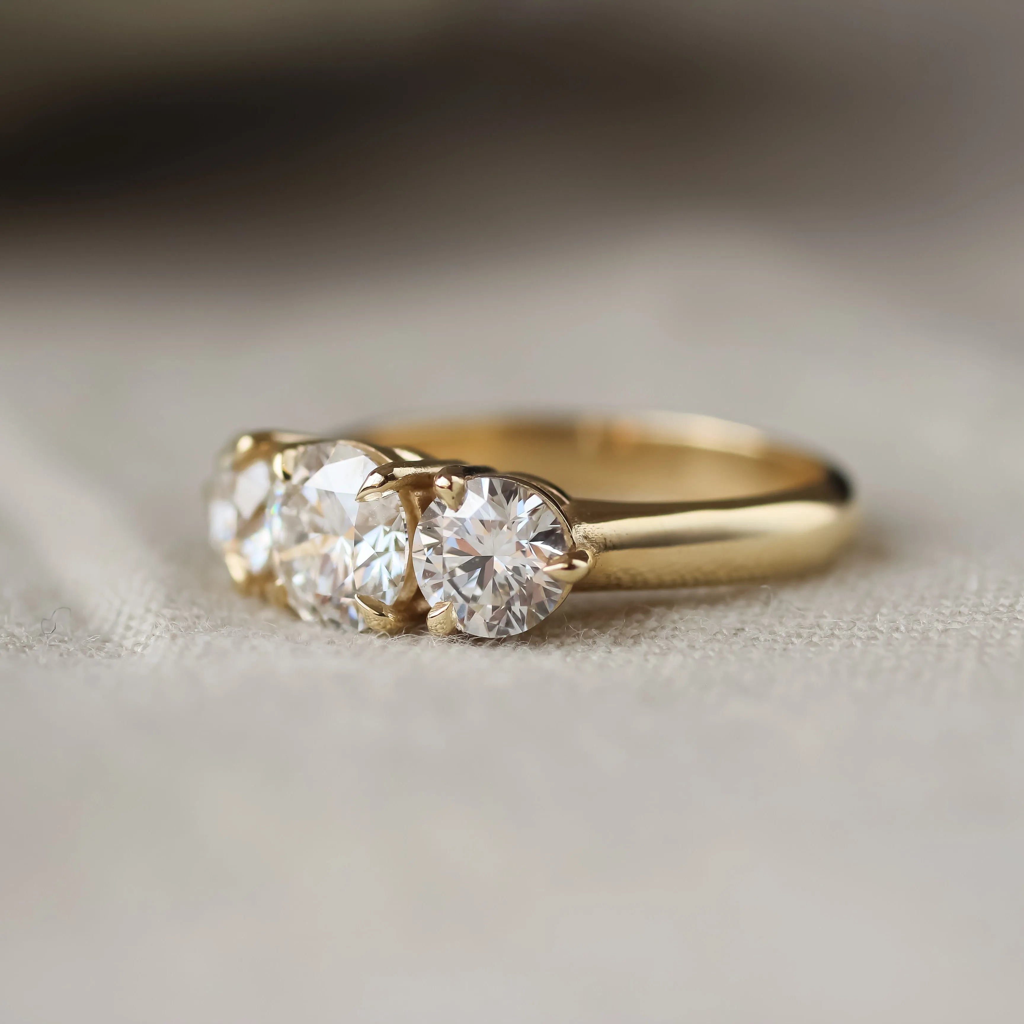 A yellow gold three-stone ring with round-cut clear gemstones, resting on a soft beige fabric surface.
