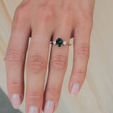 A close-up of a hand wearing a gold ring with a central oval green and teal sapphire and small clear accent stones on either side, set against a soft beige background.