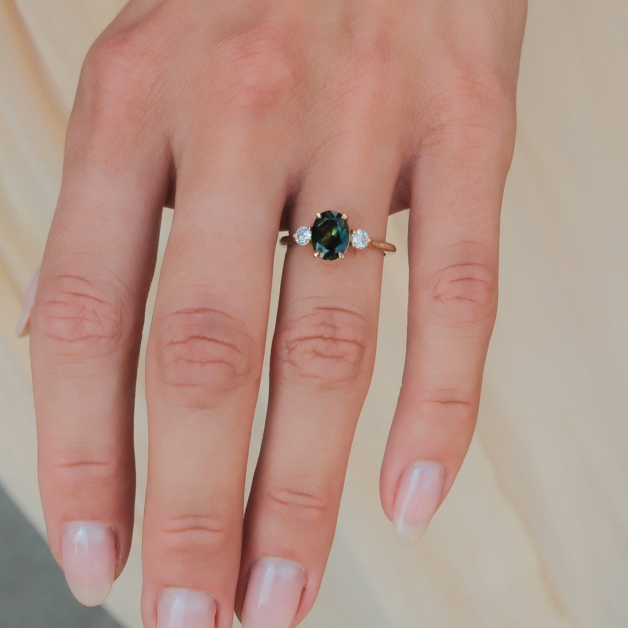 A close-up of a hand wearing a gold ring with a central oval green and teal sapphire and small clear accent stones on either side, set against a soft beige background.