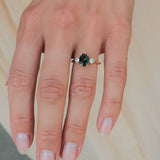 A close-up of a hand wearing a gold ring with a central oval green and teal sapphire and small clear accent stones on either side, set against a soft beige background.