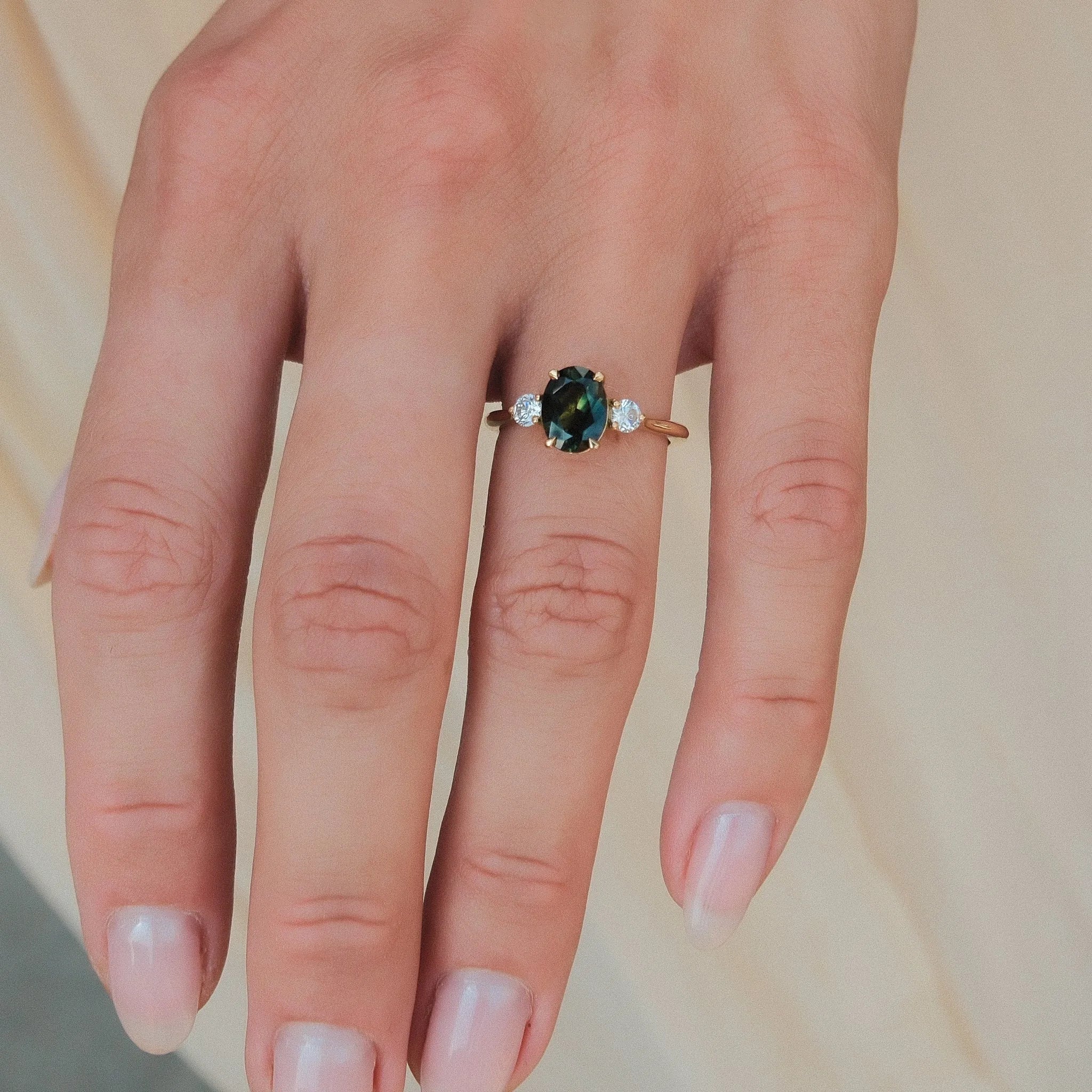 A close-up of a hand wearing a gold ring with a central oval green and teal sapphire and small clear accent stones on either side, set against a soft beige background.