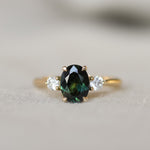 A yellow gold three-stone ring featuring an oval dark green sapphire in the center, flanked by two round clear accent stones, shown on a soft neutral background.